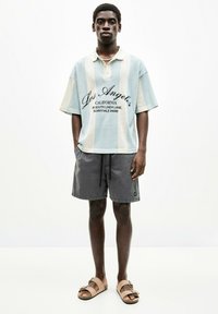 Hellblau- und cremefarbene gestreifte Kurzarm-Poloshirt mit schwarzem Druck, kombiniert mit grauen Shorts und braunen Sandalen. Lässiges Sommeroutfit.