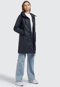 Chaqueta impermeable en color marino con capucha, de mangas largas, bolsillos laterales con cremallera y un cordón ajustable en la cintura, combinada con unos vaqueros azul claro.