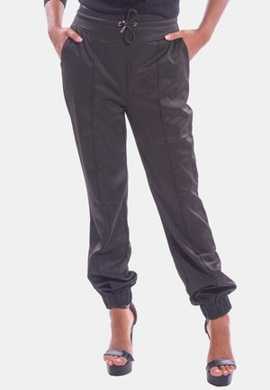Femme portant un pantalon de jogging noir avec des détails de couture devant et des poignets élastiques, associé à des sandales à talons noirs, mains dans les poches.