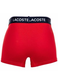 Röda bomullboxershorts med en marinblå midja med vit "LACOSTE"-logotyp. Tyget verkar slätt med en välsittande design.