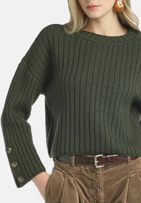 Ciemnozielony sweter z prążkowanego dzianiny z długimi rękawami, ozdobiony guzikami na mankietach. Noszony z brązowym paskiem z sztruksu.