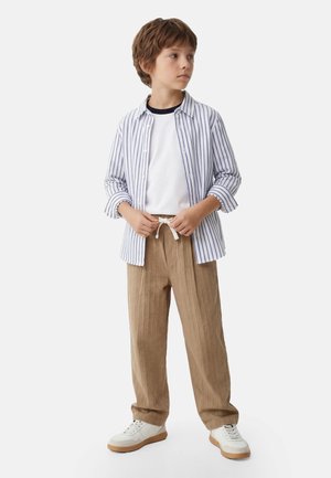 Ragazzo che indossa pantaloni beige con coulisse, sneakers bianche, t-shirt bianca e camicia aperta a righe blu, in piedi e guarda di lato.