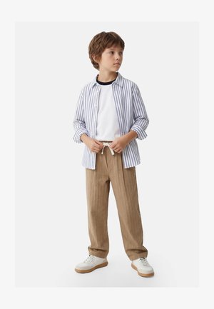 Garçon portant un pantalon beige à cordon, des baskets blanches, un t-shirt blanc et une chemise boutonnée rayée bleue ouverte, debout et regardant sur le côté.