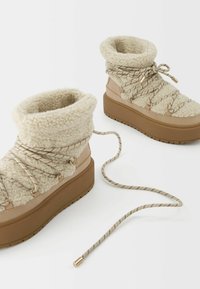 Beige støvler foret med fleece, med en tyk brun platformsål, dekoreret med snørebånd og metalaccenter. Tekstureret overdel og fleksibel form.