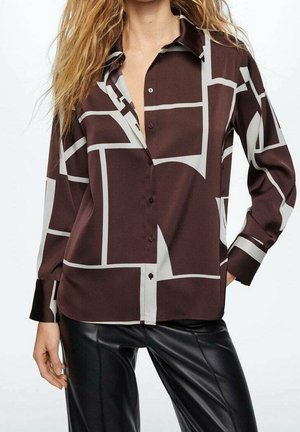 Frau trägt ein braun-beiges Hemd mit geometrischem Muster und schwarze Lederhose, steht mit einer Hand in der Hüfte.