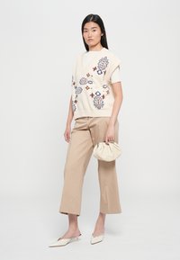 Strickweste in Creme mit farbenfroher Blumenstickerei, kombiniert mit beigen weiten Hosen und einer Creme-Clutch, dazu spitze Mules in Creme.