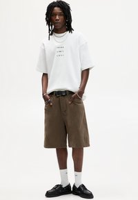 Witte oversized t-shirt met de tekst "DENK BEPERKINGLOOS", bruine corduroy shorts, zwarte riem, zwarte schoenen en witte sokken.