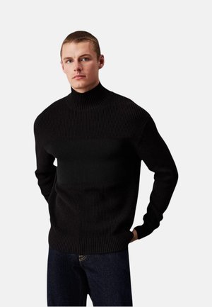 Giovane uomo con capelli corti che indossa un maglione a collo alto nero a coste e jeans scuri su uno sfondo chiaro e uniforme.