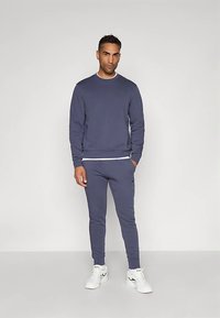 Άντρας στέκεται φορώντας ένα ταιριαστό μπλε φούτερ και φόρμες με λευκά αθλητικά παπούτσια, μπροστά από έναν απλό ανοιχτό φόντο.