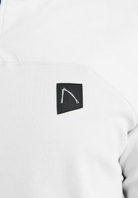 Hvid beklædning med et tekstureret, blødt stof og en sort lap, der viser et stiliseret logo med hvid stikning på venstre side.