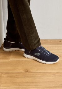 Marinblå mesh-sneakers med vita sulor, med elastiska snören och en logo på sidan. Bärs med olivgröna manchesterbyxor.