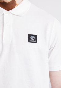 Baltas polo krekls, izgatavots no teksturēta kokvilnas, ar melnu kvadrātveida uzšuvumu, kurā attēlots koka logo un uzraksts "Timberland" baltā krāsā. Divu pogu apkakle.