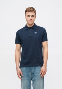 Ναυτικό πόλο μπλουζάκι από βαμβάκι, με γιακά, κοντά μανίκια και μικρό λογότυπο στο αριστερό μέρος. Συνδυάζεται με ανοιχτό μπλε τζιν.