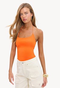 Jeune femme aux longs cheveux blonds portant un haut dos nu orange, un pantalon cargo blanc, des boucles d'oreilles créoles en or et un bracelet large en or.