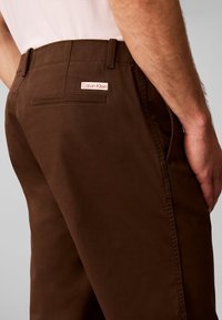 Άνδρας φοράει καφέ παντελόνι Calvin Klein, δείχνοντας την πίσω τσέπη και τη ραφή στο πλάι, με το χέρι να ακουμπά στο πλάι του.