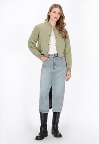 Femeie îmbrăcată cu o jachetă matlasată verde măsliniu, un top alb, o fustă midi din denim albastru deschis cu o crestătură frontală și cizme negre până la genunchi, stând cu o mână în buzunar.