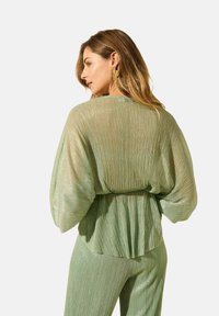 Haut vert, transparent, plissé avec des manches chauve-souris, taille doucement froncée et finition texturée, associé à un pantalon plissé assorti.