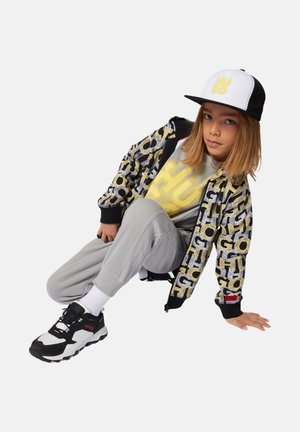 Kind dat een zwart-wit petje draagt, een patroonjas, een geel shirt, een grijze joggingbroek en zwart-witte sneakers, zittend op de vloer tegen een witte achtergrond.