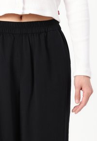 Black, elastic-waist pants made of smooth fabric. The top section is cropped white ribbed material with buttons, exposing midriff.