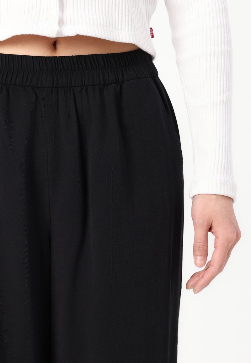 Schwarze, elastische Hosen mit einem glatten Stoffbund. Der obere Teil besteht aus einem kurzen weißen Rippmaterial mit Knöpfen, das den Bauch freilegt.
