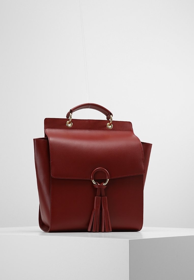 Sac à dos en cuir rouge avec un design structuré, une poignée arrondie, une fermeture à rabat, des accents métalliques et un détail décoratif en pampille.