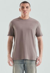Hellbraunes Kurzarm-T-Shirt aus weicher Baumwolle mit rundem Halsausschnitt und einem kleinen Logodetail auf der Brust. Kombiniert mit ausgebleichten Jeans.