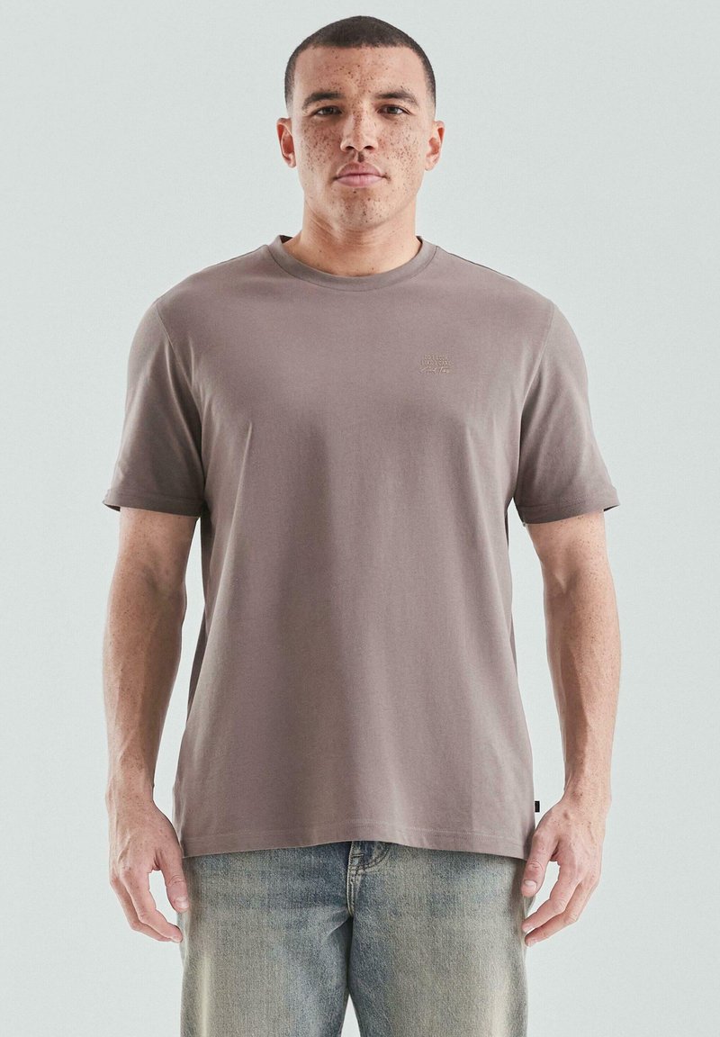 Hellbraunes Kurzarm-T-Shirt aus weicher Baumwolle mit rundem Halsausschnitt und einem kleinen Logodetail auf der Brust. Kombiniert mit ausgebleichten Jeans.
