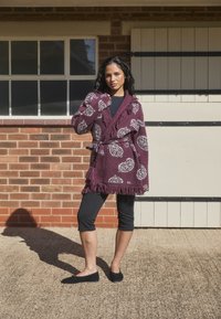 Cárdigan burdeos con estampado y dobladillo con flecos, usado sobre una blusa negra y leggings. Zapatos negros planos completan el atuendo. Pared de ladrillo al fondo.