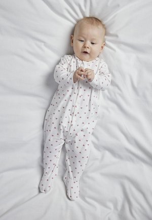 Bébi fehér, apró piros szívecskékkel díszített lábfejes pizsamában, fehér ágyneműn fekszik, lefelé nézve, miközben a kezét érinti.