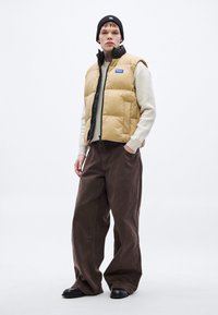 Beežikarva pufferi vest musta luku jaamu, kantud kreemja kampsuni ja pruuni manšestrpükstega. Must moekas müts ja mustad kingad lõpetavad komplekti.