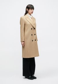 Μπεζ παλτό διπλής όψης με μαύρα κουμπιά, μακριά μανίκια και ραμμένη εφαρμογή. Συνδυασμένο με μαύρο παντελόνι φαρδύ από το μπούτι και μαύρα μποτάκια αστραγάλου.