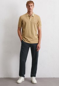 Beżowa koszulka polo z kołnierzykiem, jasny akcent na dekolcie, noszona z ciemnymi spodniami i białymi sneakersami. Model stoi z rękami w kieszeniach.