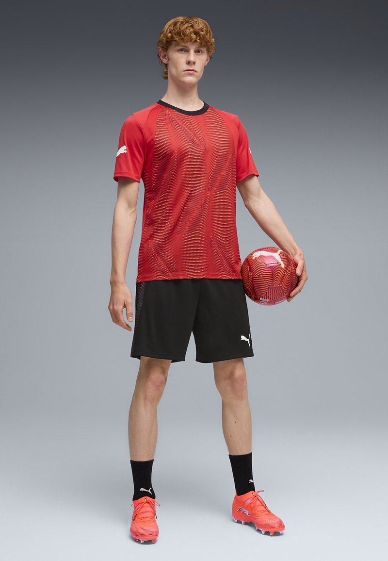 Camiseta deportiva roja con patrón texturizado, detalles en negro, pantalones cortos negros, tacos naranja brillante, sosteniendo un balón de fútbol colorido.