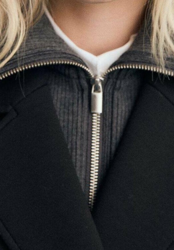 Gros plan d'une personne portant un manteau noir par-dessus un pull gris à fermeture éclair avec un col blanc visible en dessous.