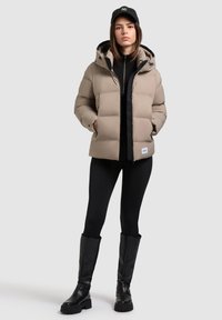 Chaqueta de plumas beige con capucha, cierre de cremallera y detalles negros, combinada con leggings negros y botas altas negras.