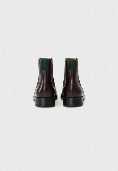 Botas de tobillo de cuero marrón con un acabado suave, suela de goma negra y tirador trasero de ante verde. Diseño minimalista y paneles laterales elásticos.