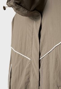 Gros plan sur une veste taupe avec un détail de passepoil blanc et une capuche à cordon, montrant la texture du tissu et la couture avant.