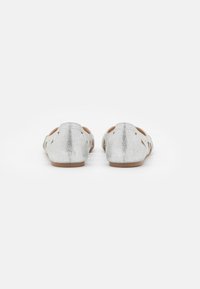 Un par de zapatos planos plateados con purpurina y detalles troquelados, vistos desde atrás sobre un fondo blanco liso.