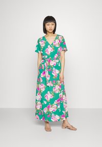 Vestido maxi florido verde com mangas curtas, decote em V, cintura com laço, apresentando grandes flores rosas e brancas num fundo vibrante.