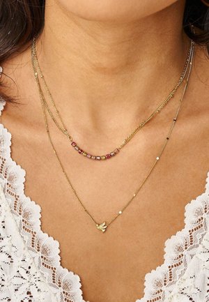 Dos delicados collares de oro en el cuello de una mujer, uno con cuentas multicolores y el otro con un pequeño colgante de pájaro, sobre una blusa blanca de encaje.