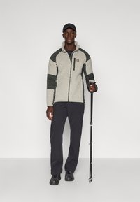 Grijze en groene ritsfleecejas met hoge kraag, gecombineerd met donkere broeken en zwarte schoenen, terwijl hij een wandelstok vasthoudt.