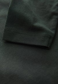 Ciemnozielona bawełniana tkanina o gładkiej fakturze, z złożonym mankietem rękawa i delikatnymi detalami szwów.