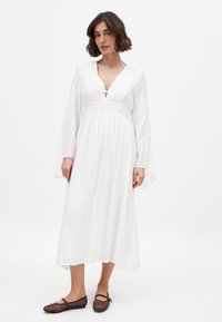 Hvid maxi kjole med lange klokkeærmer, med en snøre lukning ved halsen og en elastisk talje. Sammen med brune ballet flade sko.