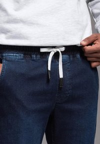 Joggers de mezclilla en azul oscuro con cintura elástica, que cuentan con un cordón blanco y bolsillos laterales. Textura suave y diseño ajustado.