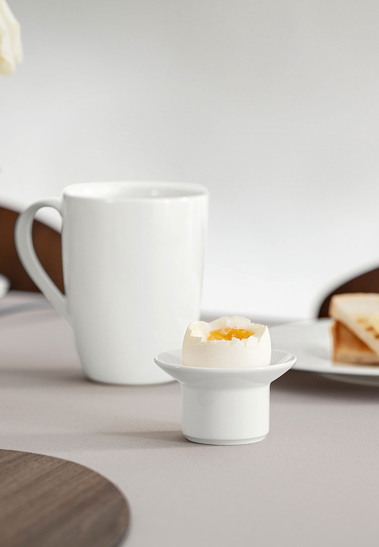 Miękko gotowane jajko w białym kubku na jajka, biały kubek za nim oraz tostowany sandwich na białym talerzu na jasnoszarym stole.