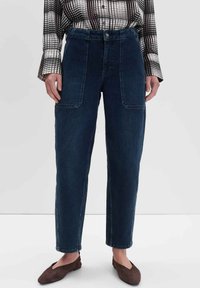 Donkerblauwe denimbroek met een hoge taille, rechte pijpen en twee voorzakken. Ggedragen met een zwart-wit geblokt overhemd.
