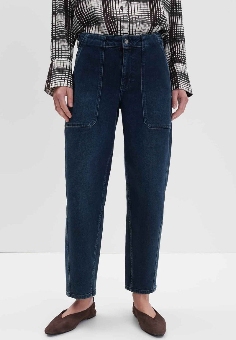 Donkerblauwe denimbroek met een hoge taille, rechte pijpen en twee voorzakken. Ggedragen met een zwart-wit geblokt overhemd.