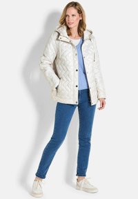 Gepolsterte, hellbeige Jacke mit Kapuze, ausgestattet mit Druckknöpfen, Seitentaschen und einem Rautenmuster; getragen über einem blauen Oberteil und Jeans.