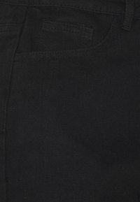 Tecido de denim preto apresentando uma visão em close dos grãos e da textura, com costuras visíveis e detalhes dos bolsos ao lado.