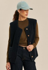 Marineblauwe fleece vest met een gestructureerde afwerking, ronde halslijn en houten knopen. Gemaakt over een bruine top, gecombineerd met donkere jeans.
